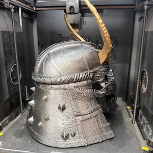 May include: A metallic silver 3D-printed helmet with a textured finish, featuring a prominent gold-colored curved horn on top and small pyramid-shaped spikes along the back. The helmet has a segmented design with visible layers and a dark interior.