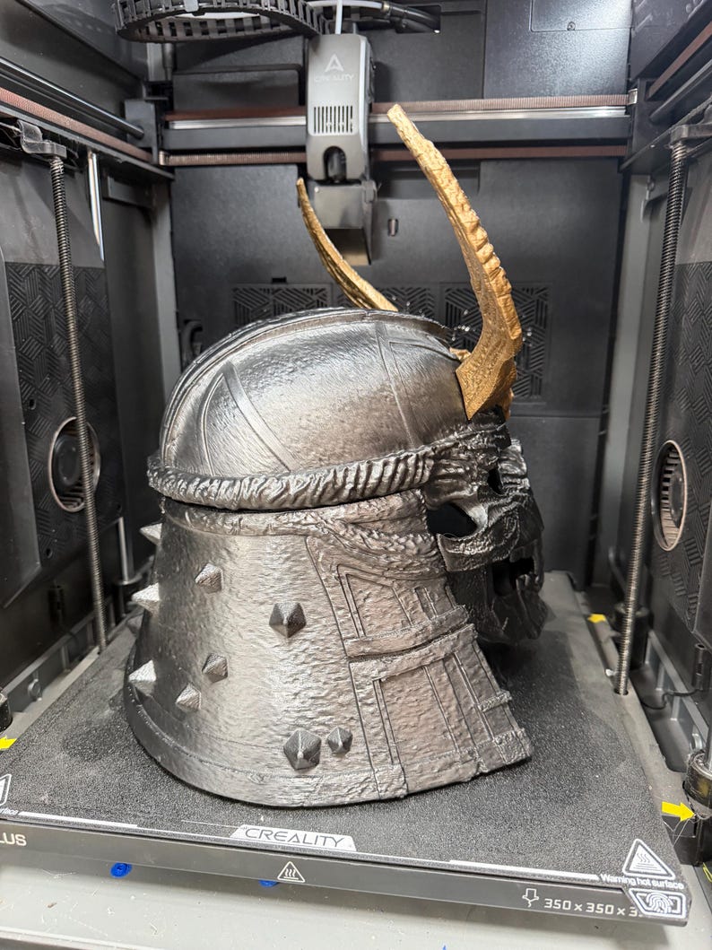 May include: A metallic silver 3D-printed helmet with a textured finish, featuring a prominent gold-colored curved horn on top and small pyramid-shaped spikes along the back. The helmet has a segmented design with visible layers and a dark interior.