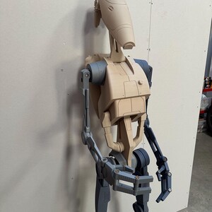 May include: A partially assembled, unpainted, beige plastic model of a Star Wars Battle Droid. The droid is standing upright with its arms and legs partially assembled. The droid's head is attached to its body, and its arms are extended out to the sides. The droid's legs are bent at the knees, and its feet are flat on the ground.