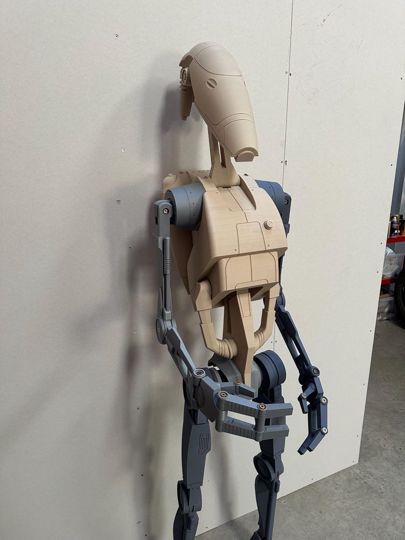 May include: A partially assembled, unpainted, beige plastic model of a Star Wars Battle Droid. The droid is standing upright with its arms and legs partially assembled. The droid's head is attached to its body, and its arms are extended out to the sides. The droid's legs are bent at the knees, and its feet are flat on the ground.