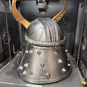 May include: A 3D printed helmet with a metallic silver finish, featuring a textured surface, a series of small pyramid-shaped studs around the base, and two large, curved, gold-colored horns extending upward from the top of the dome.