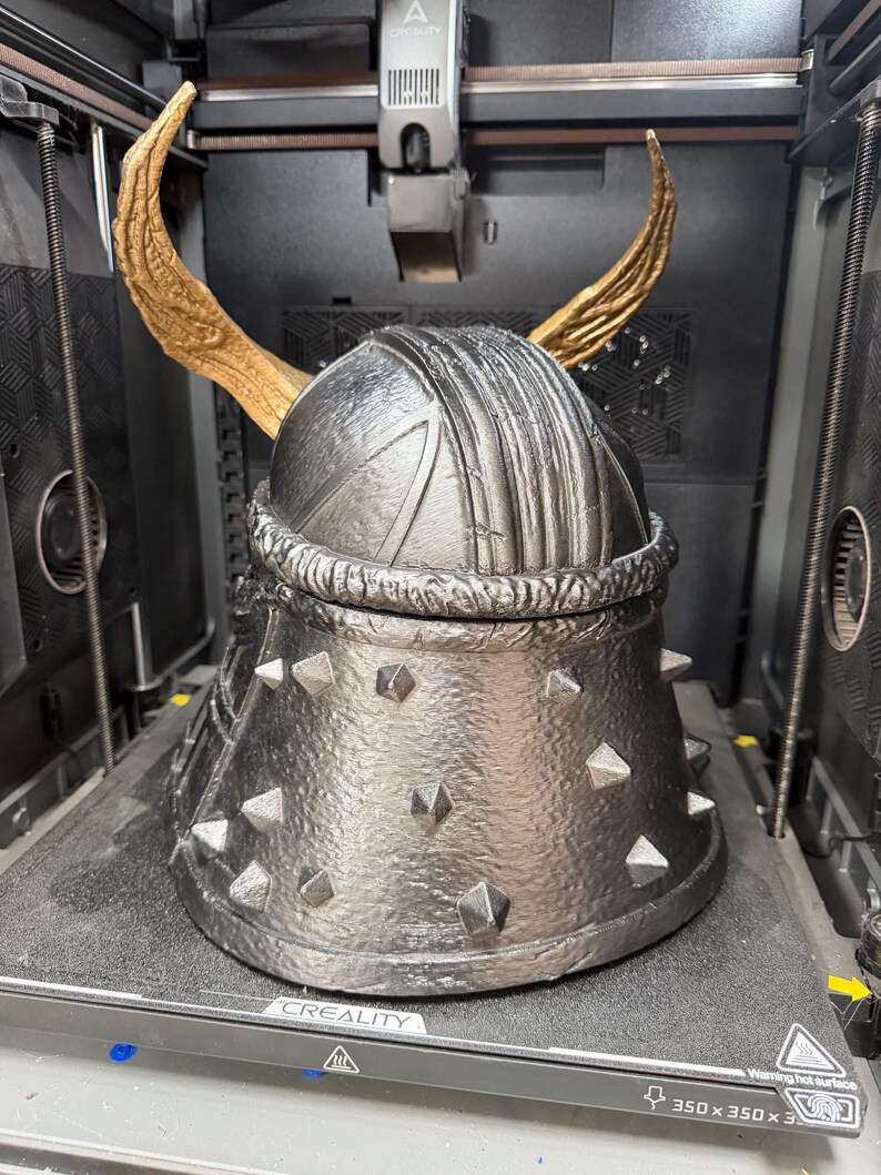 May include: A 3D printed helmet with a metallic silver finish, featuring a textured surface, a series of small pyramid-shaped studs around the base, and two large, curved, gold-colored horns extending upward from the top of the dome.