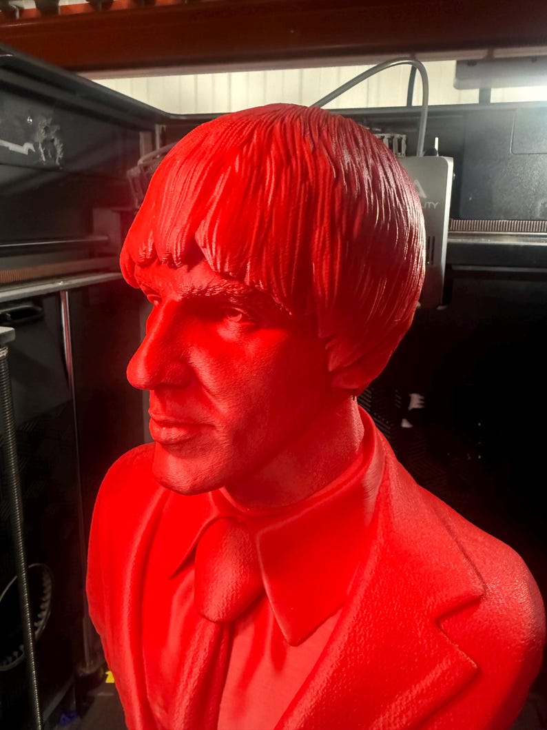 May include: A solid red bust of a man with a distinctive hairstyle and suit. The sculpture is a side profile, showcasing the man's features. The bust is likely a decorative item or a piece of art.