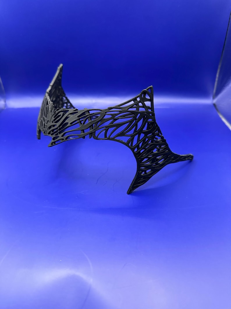 May include: A black headpiece with a web-like, open design. The headpiece has a pointed, angular shape. The object is set against a solid blue background. The intricate pattern gives it a delicate yet striking appearance.