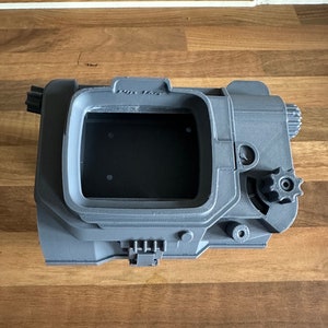 May include: A grey 3D printed model of a Pip-Boy from the Fallout video game series. The model features a screen, buttons, and a gear on the side.