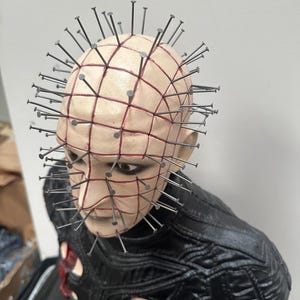May include: A detailed bust of a character with a pale complexion, a grid-like pattern of red lines, and numerous metal nails embedded in the head. The figure wears a black, textured garment with red accents. The overall aesthetic is dark and gothic.