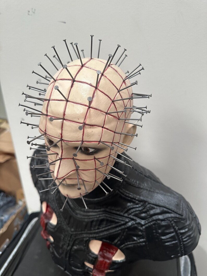 May include: A detailed bust of a character with a pale complexion, a grid-like pattern of red lines, and numerous metal nails embedded in the head. The figure wears a black, textured garment with red accents. The overall aesthetic is dark and gothic.