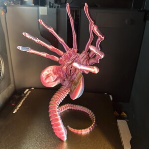 Op de afbeelding: Een 3D-geprint model van een buitenaards wezen met een lange, gesegmenteerde staart en meerdere ledematen. Het model is geprint in een roze en paarse kleurschema en staat op een zwart oppervlak.