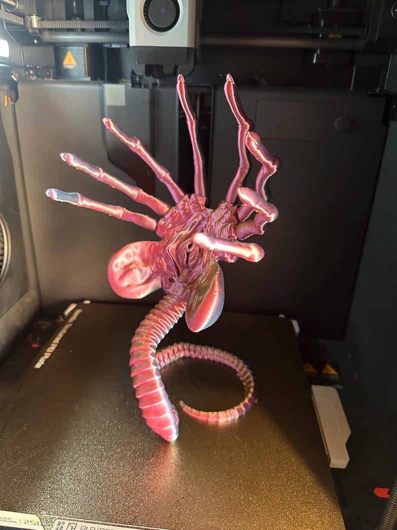 Op de afbeelding: Een 3D-geprint model van een buitenaards wezen met een lange, gesegmenteerde staart en meerdere ledematen. Het model is geprint in een roze en paarse kleurschema en staat op een zwart oppervlak.