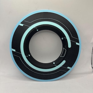 May include: A circular black and light blue object with a futuristic design. The object has a light blue outer ring, a black inner ring, and a series of light blue and white lines and shapes. The object is likely a prop or decorative item.