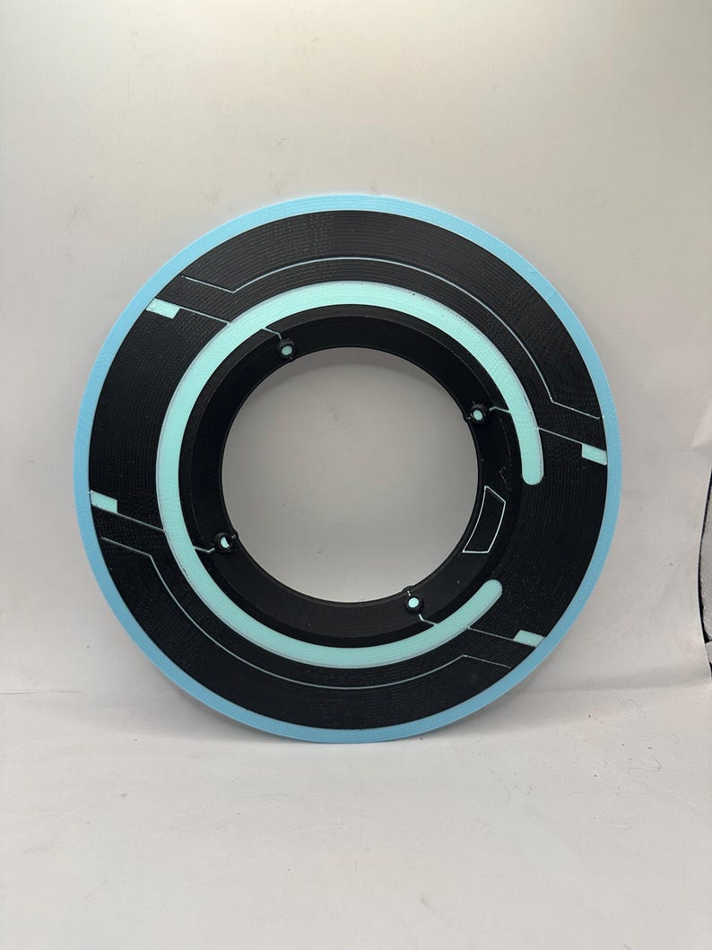 May include: A circular black and light blue object with a futuristic design. The object has a light blue outer ring, a black inner ring, and a series of light blue and white lines and shapes. The object is likely a prop or decorative item.