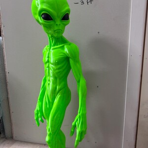Pode incluir: Uma figura alien&iacute;gena verde neon brilhante com uma cabe&ccedil;a grande, membros alongados e olhos ovais escuros. A figura est&aacute; em p&eacute; e apresenta o texto -3 ft escrito na parede atr&aacute;s, indicando uma altura de aproximadamente 91 cm.