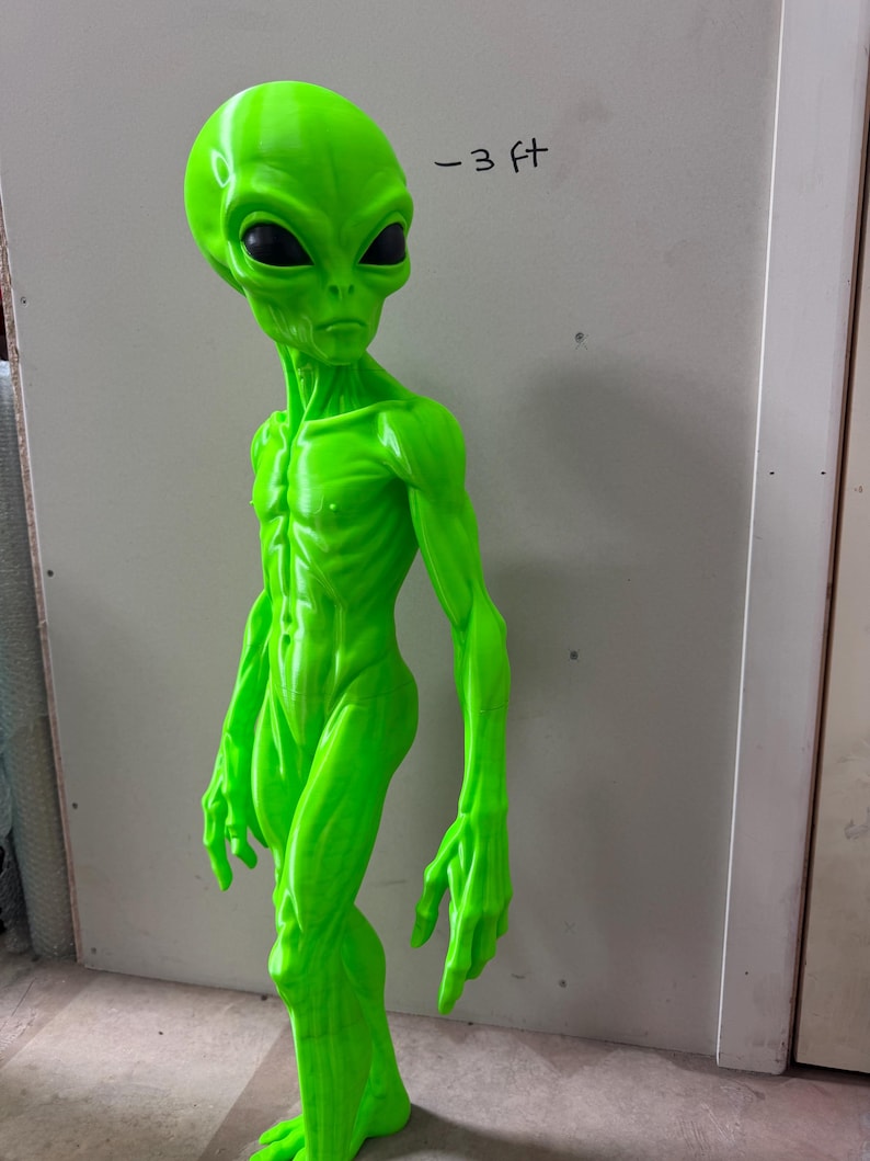 Pode incluir: Uma figura alien&iacute;gena verde neon brilhante com uma cabe&ccedil;a grande, membros alongados e olhos ovais escuros. A figura est&aacute; em p&eacute; e apresenta o texto -3 ft escrito na parede atr&aacute;s, indicando uma altura de aproximadamente 91 cm.