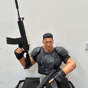 May include: A detailed bust of a muscular figure in a black tactical outfit, holding two black rifles. The figure has short, dark hair and a stern expression. The bust is set against a plain white background.