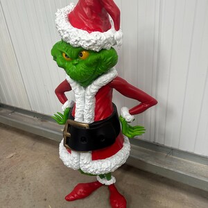 May include: A green Grinch figure wearing a red Santa suit with white trim and a matching hat. The figure has yellow eyes and is posed with arms akimbo, a black belt, and red shoes. A festive Christmas decoration.