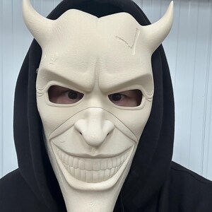 May include: A cream-colored mask with curved horns and a wide, unsettling grin. The mask has eye openings and is worn over a black hooded sweatshirt. The mask's surface has a textured appearance and a crack on the forehead.
