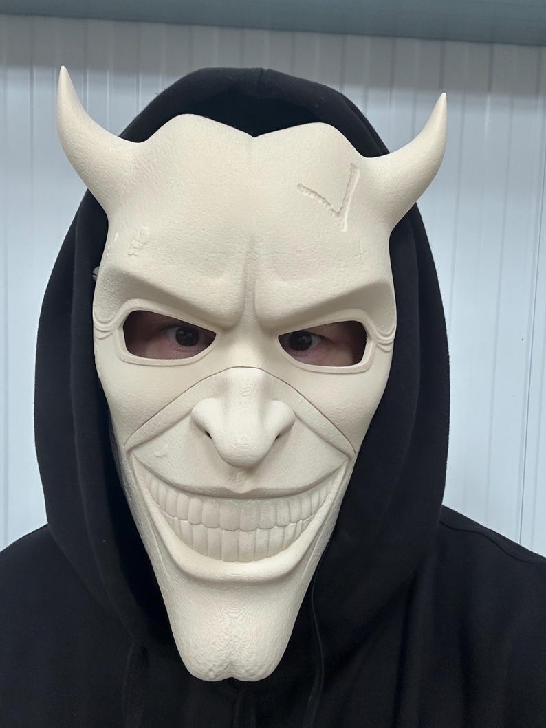 May include: A cream-colored mask with curved horns and a wide, unsettling grin. The mask has eye openings and is worn over a black hooded sweatshirt. The mask's surface has a textured appearance and a crack on the forehead.