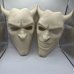 May include: Two off-white, horned masks with menacing expressions. One mask has a neutral expression, while the other has a wide, toothy grin. Both masks feature eye holes, a nose, and a chin.