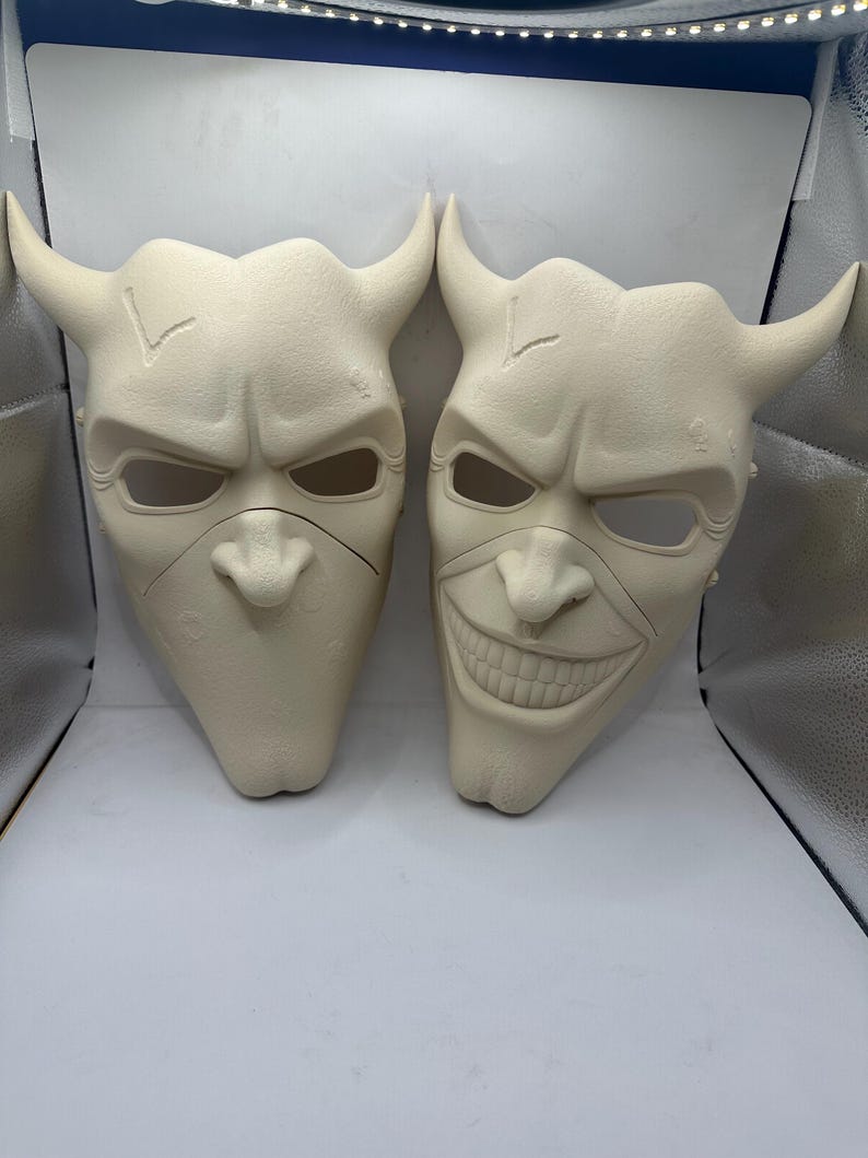 May include: Two off-white, horned masks with menacing expressions. One mask has a neutral expression, while the other has a wide, toothy grin. Both masks feature eye holes, a nose, and a chin.