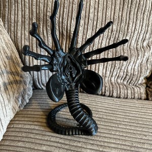 Op de afbeelding: Zwarte plastic figuur van een buitenaards wezen met meerdere poten en een lange staart. Het wezen heeft een gedetailleerd, gesegmenteerd lichaam en een grote, open mond.