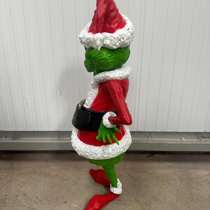 May include: A green figure in a red Santa suit with a matching hat and white trim. The figure has a black belt and red shoes. The figure is a Christmas decoration.