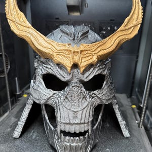 May include: A silver-colored skull-shaped helmet with a textured, weathered finish and prominent teeth. The helmet features large, curved, gold-colored horns extending from the forehead area. The word CREALITY is printed on the base.