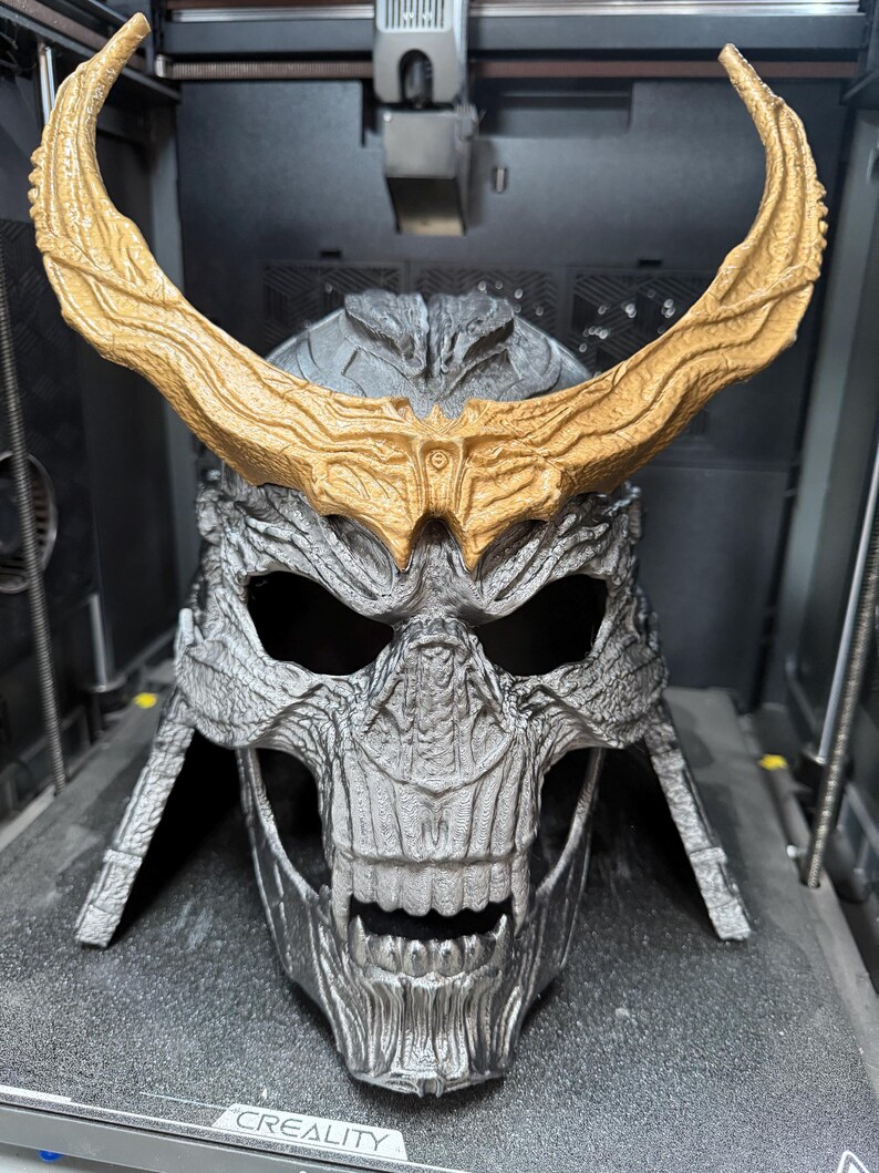 May include: A silver-colored skull-shaped helmet with a textured, weathered finish and prominent teeth. The helmet features large, curved, gold-colored horns extending from the forehead area. The word CREALITY is printed on the base.