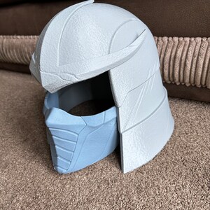 May include: A white plastic 3D printed helmet with a blue plastic face mask. The helmet has a textured surface and a raised design on the top. The face mask has a textured surface and a raised design on the front.