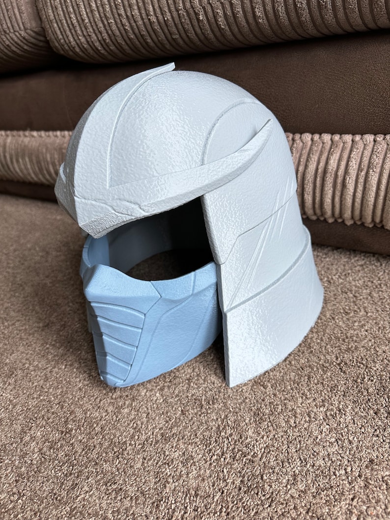 May include: A white plastic 3D printed helmet with a blue plastic face mask. The helmet has a textured surface and a raised design on the top. The face mask has a textured surface and a raised design on the front.