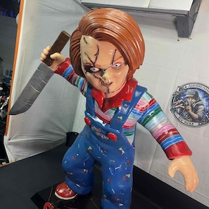 May include: A Chucky doll figurine with a knife. The doll has red hair, a stitched face, and is wearing blue overalls with red shoes. The base reads "CHUCKY".
