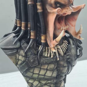Puede incluir: Busto detallado de un alien&iacute;gena Predator, con una cabeza texturizada marr&oacute;n y beige, rastas y una boca abierta que revela dientes afilados. El busto incluye una pieza de pecho detallada con un patr&oacute;n de diamantes y una base gris.