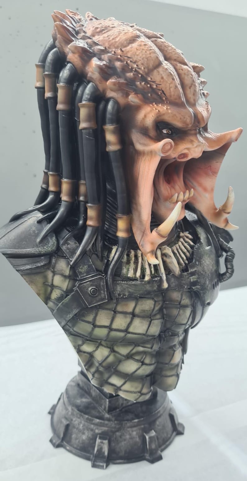 Puede incluir: Busto detallado de un alien&iacute;gena Predator, con una cabeza texturizada marr&oacute;n y beige, rastas y una boca abierta que revela dientes afilados. El busto incluye una pieza de pecho detallada con un patr&oacute;n de diamantes y una base gris.