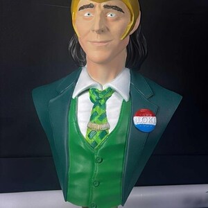 May include: A bust of a character wearing a gold horned helmet, green suit, and a green tie. The bust has a pin that says "LOKI". The base of the bust is gold with the word "LOKI" on it.