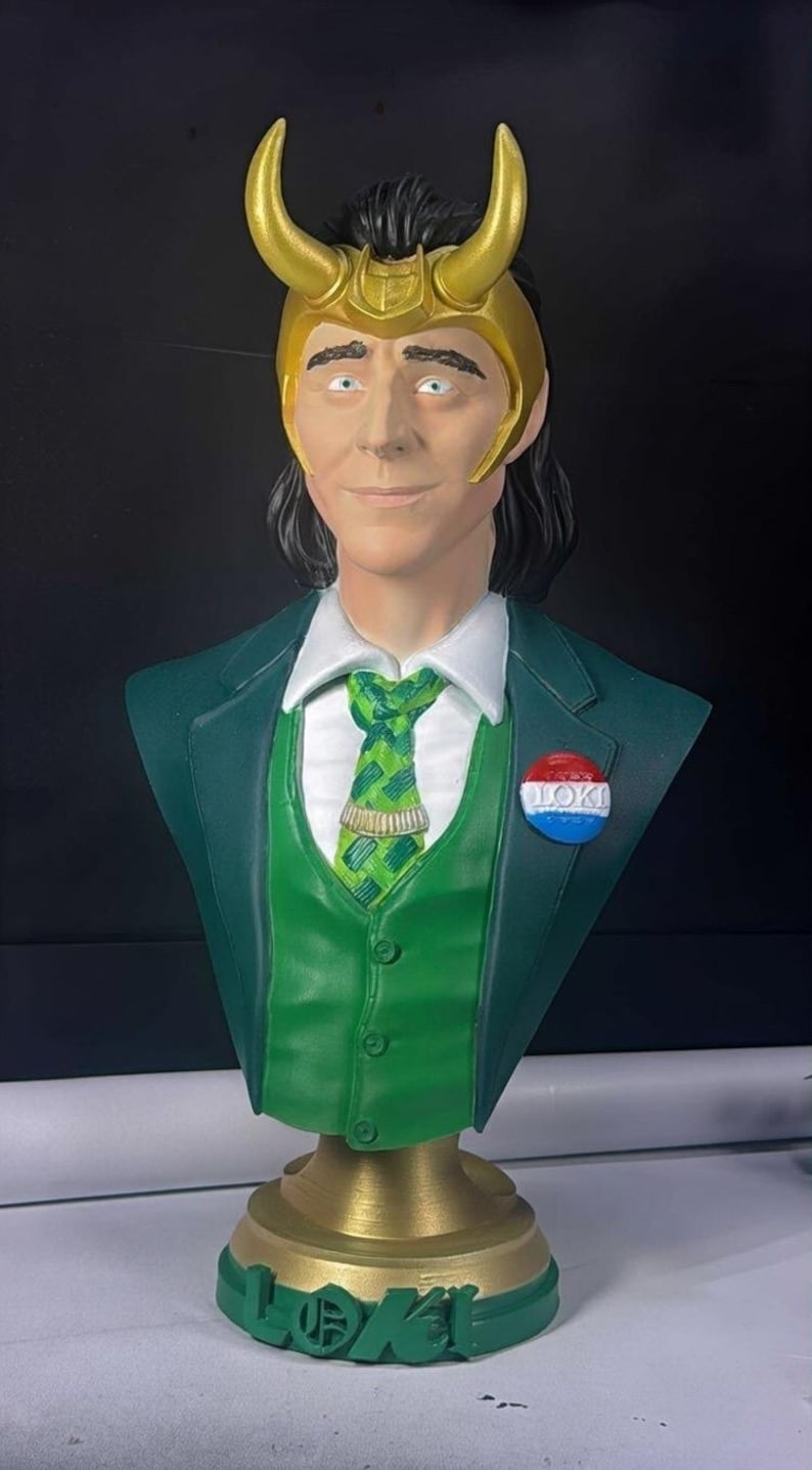 May include: A bust of a character wearing a gold horned helmet, green suit, and a green tie. The bust has a pin that says "LOKI". The base of the bust is gold with the word "LOKI" on it.