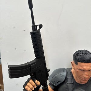 May include: A black rifle with a silencer is held by a person wearing a black shirt and shoulder armor. The rifle has a magazine and a detailed design. The person's face is visible, with short black hair and a serious expression.