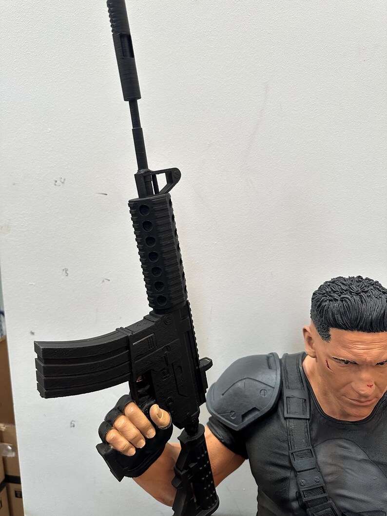 May include: A black rifle with a silencer is held by a person wearing a black shirt and shoulder armor. The rifle has a magazine and a detailed design. The person's face is visible, with short black hair and a serious expression.