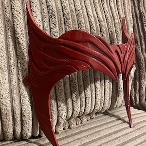 May include: A red plastic headband with a detailed, intricate design. The headband is curved and has a pointed shape on each end.