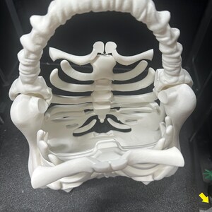 May include: A 3D-printed, off-white skeleton-shaped basket. The basket features a rib cage design forming the body, with a curved handle made of vertebrae. The base is a flat surface, and the overall structure is detailed.