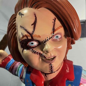May include: A detailed, close-up of a Chucky doll. The doll has red hair, stitched facial wounds, and blue eyes. It wears a blue overall with a red shirt underneath. The doll's expression is a sinister smile.