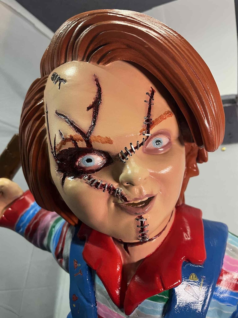 May include: A detailed, close-up of a Chucky doll. The doll has red hair, stitched facial wounds, and blue eyes. It wears a blue overall with a red shirt underneath. The doll's expression is a sinister smile.