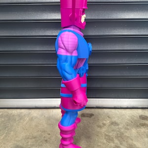 May include: A side view of a large, stylized action figure of Galactus. The figure is predominantly magenta and blue, with a rectangular head and a detailed costume. The figure is standing on a concrete surface.