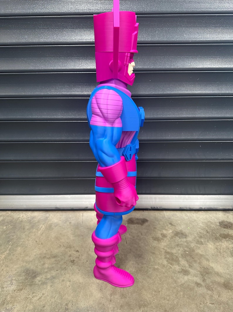 May include: A side view of a large, stylized action figure of Galactus. The figure is predominantly magenta and blue, with a rectangular head and a detailed costume. The figure is standing on a concrete surface.