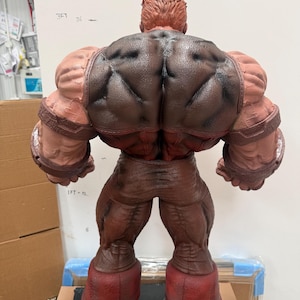 May include: A large, detailed figurine of a muscular character, viewed from the back. The figure is predominantly brown and red, with textured skin and clothing. The boots are a deep red, and the figure is posed in a standing position.