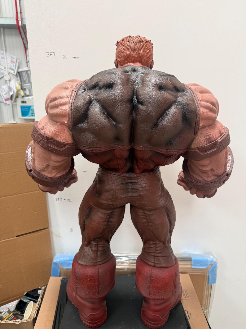 May include: A large, detailed figurine of a muscular character, viewed from the back. The figure is predominantly brown and red, with textured skin and clothing. The boots are a deep red, and the figure is posed in a standing position.