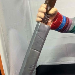 May include: A prop knife with a silver blade and a brown handle. The handle is held by a hand with a red, blue, and white striped sleeve. The hand is painted with a light skin tone.