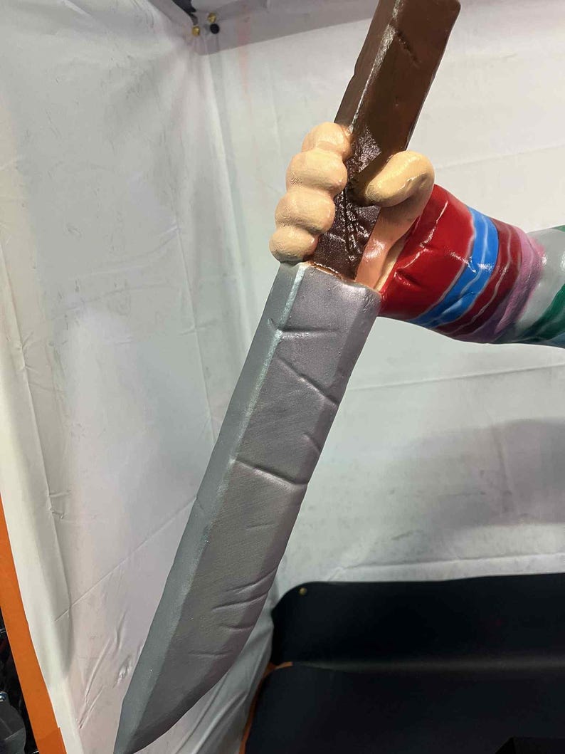 May include: A prop knife with a silver blade and a brown handle. The handle is held by a hand with a red, blue, and white striped sleeve. The hand is painted with a light skin tone.