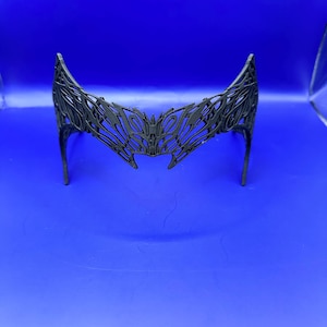 May include: A black, open-work crown or headpiece with a symmetrical design. The headpiece has a curved shape with pointed tips and a central design element. The intricate pattern suggests a decorative or costume piece, possibly for cosplay or themed events.