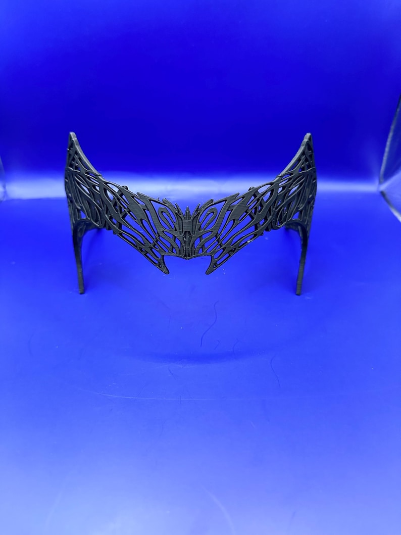 May include: A black, open-work crown or headpiece with a symmetrical design. The headpiece has a curved shape with pointed tips and a central design element. The intricate pattern suggests a decorative or costume piece, possibly for cosplay or themed events.