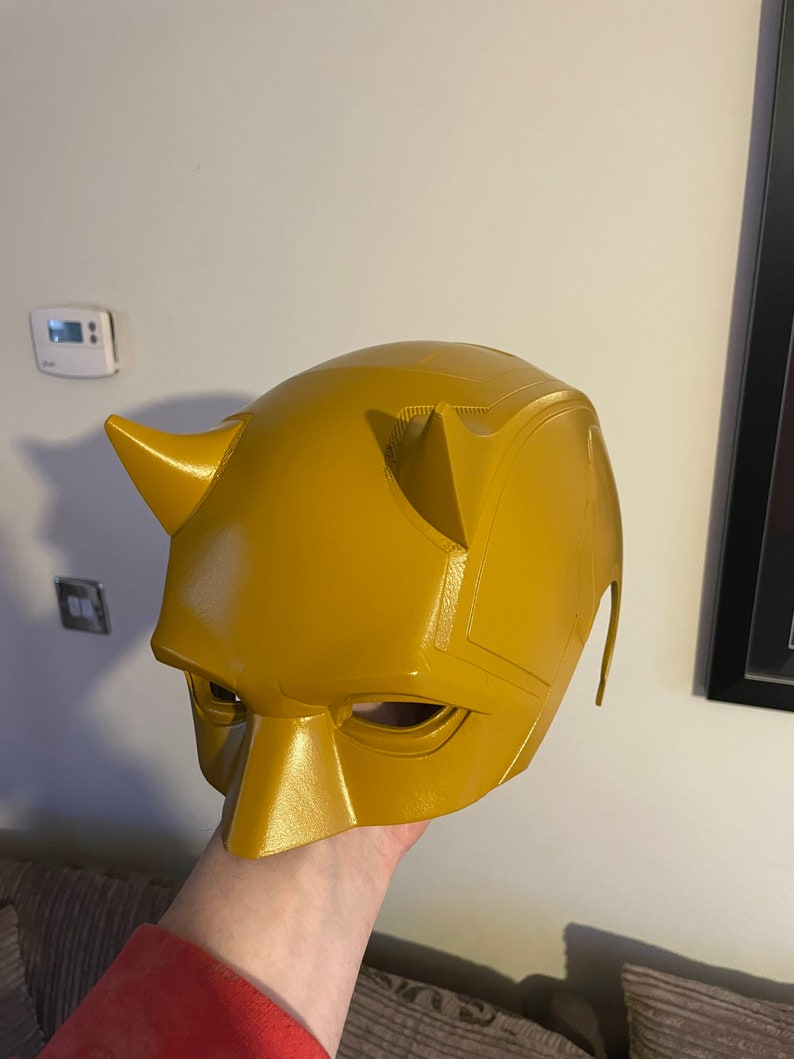 May include: A yellow plastic Daredevil mask with horns and eye holes. The mask is held in a hand.