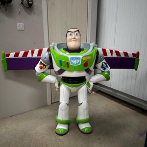May include: A large Buzz Lightyear action figure, featuring a white, green, and purple spacesuit design. The figure has a smiling face and outstretched wings with red and white stripes. The figure is standing on a concrete floor.